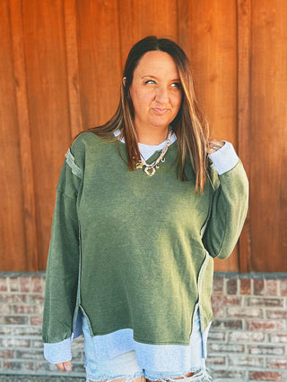 A woman wearing an oversized sage green knit sweater with light blue and white color-blocked details on the sleeves and hem, paired with denim shorts, standing in front of a wooden wall.